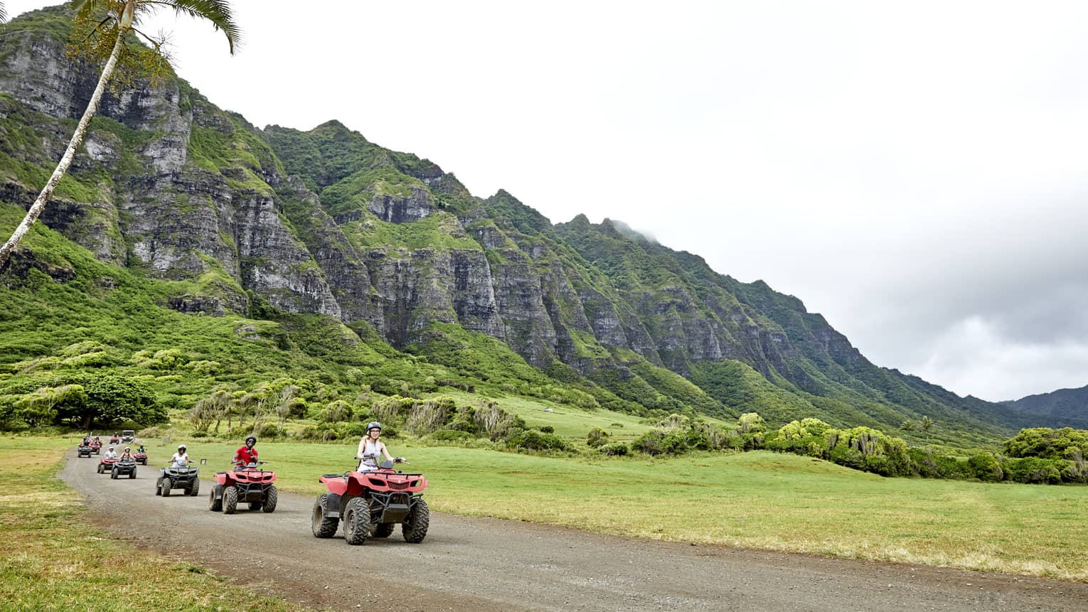 A row of ATV riders ride toward the foreground, leaving behind a verdant mountain with craggy rock rising into misty clouds.