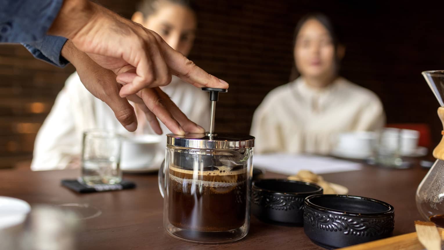 Fingers push on the piston of a small French coffee press, filtering the frothy liquid as two seated guests observe. 