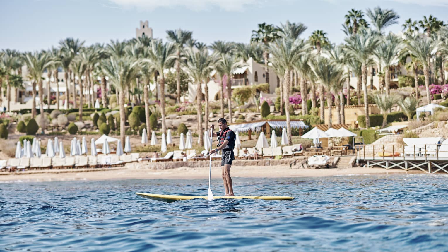A paddler stands on a board in the water in front of a beach lined with lounge chairs and palm trees and a villa beyond.