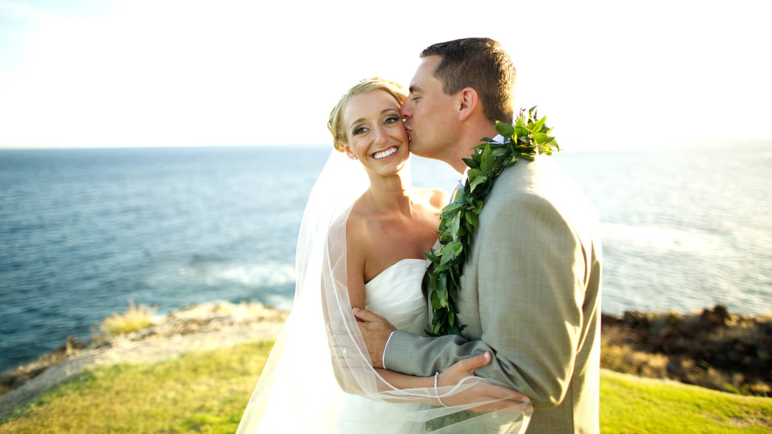 Groom kisses smiling bride on lawn of cliff overlooking ocean on sunny day
