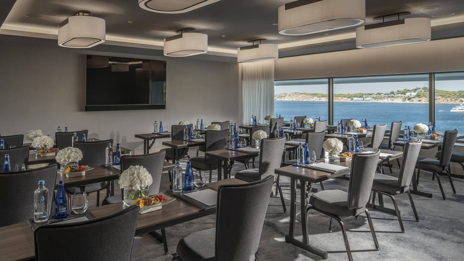 Rows of desks with water, flowers, snacks and grey chairs facing TV with ocean view