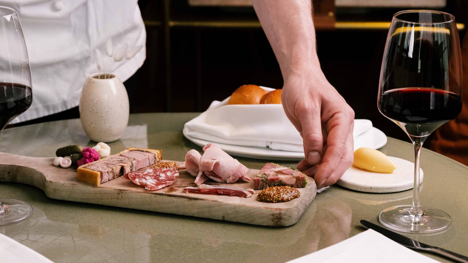 Chef places a wooden board of charcuterie on table next to glass of red wine