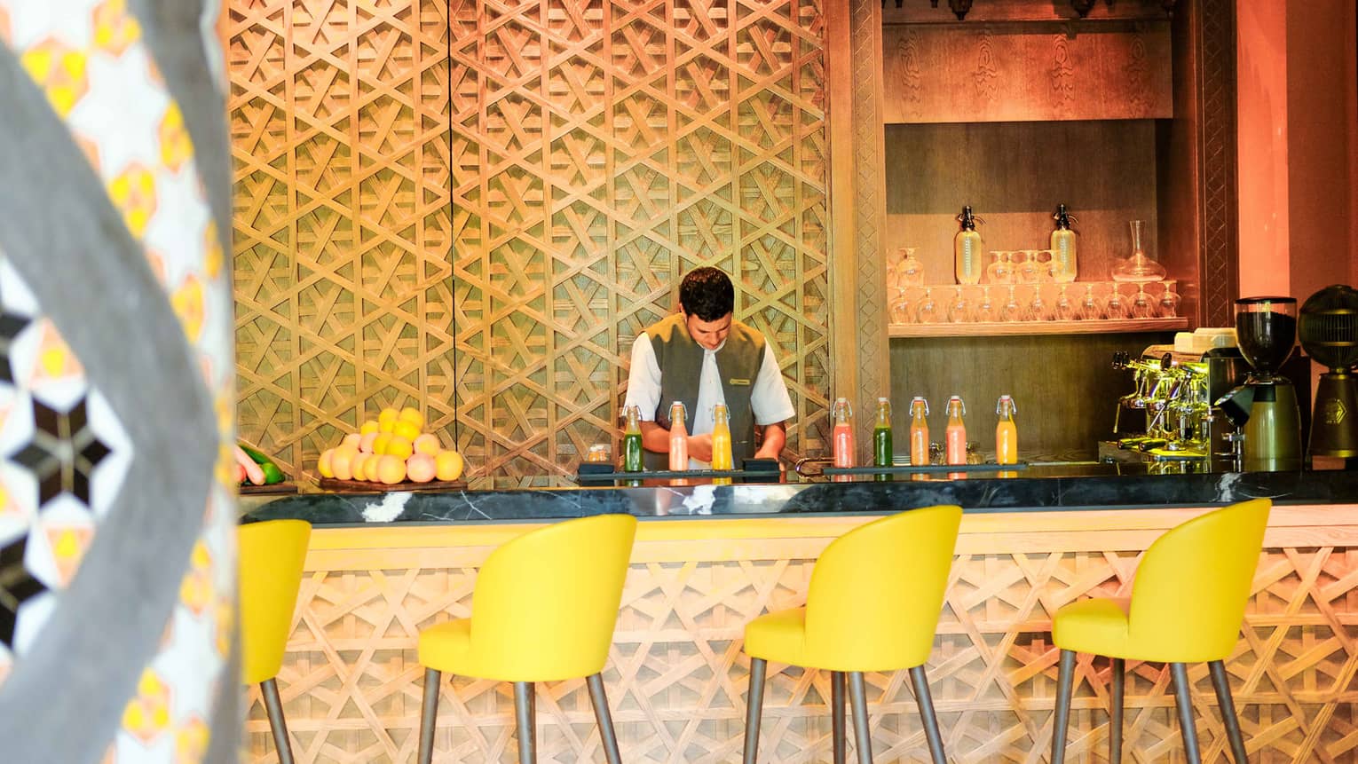 Bartender with fresh juice in glass jugs, fresh oranges on decorative carved bar lined with yellow stools