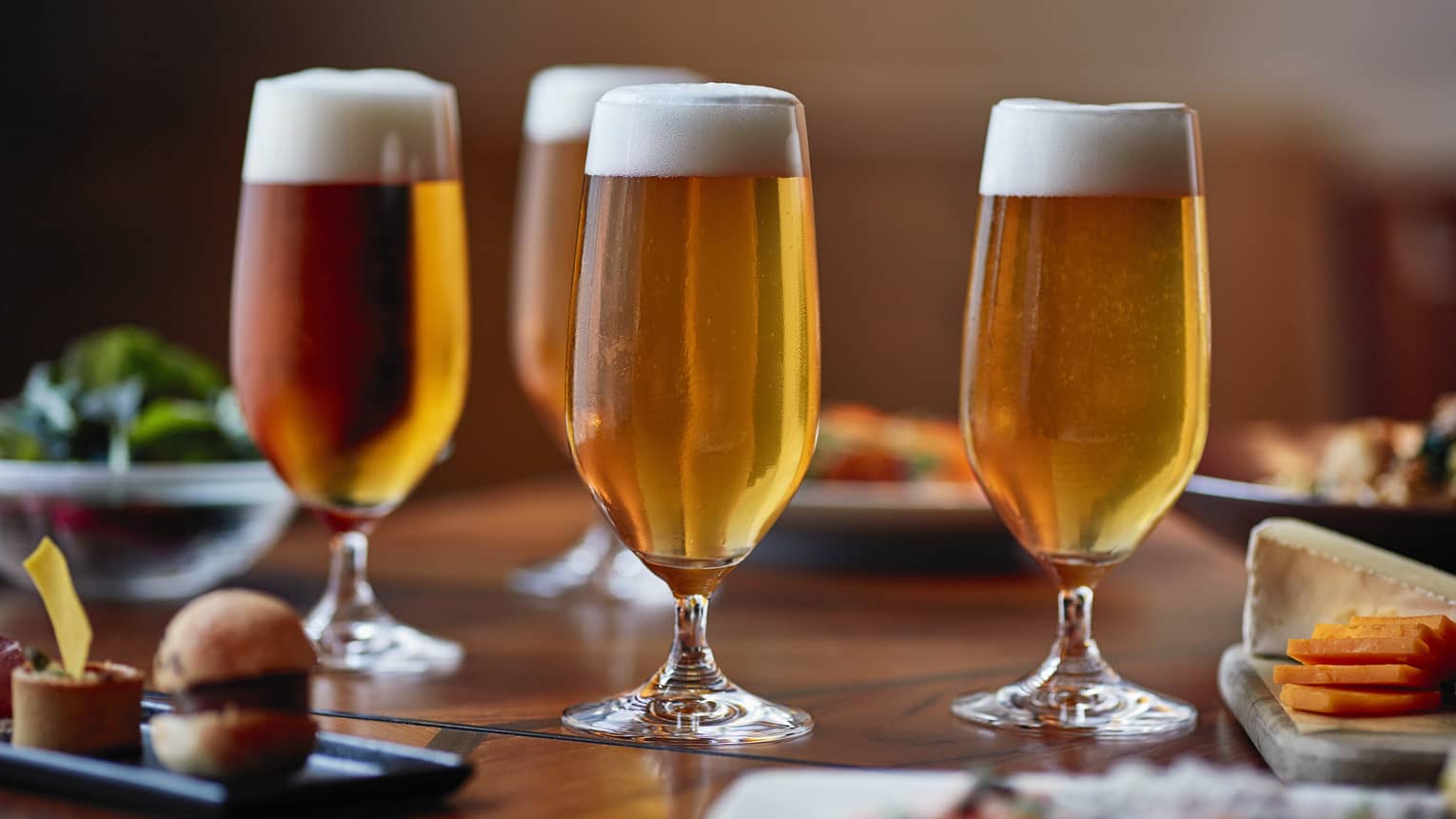 Four tall beer glasses filled with beer on a table with cheese, crackers and appetizers