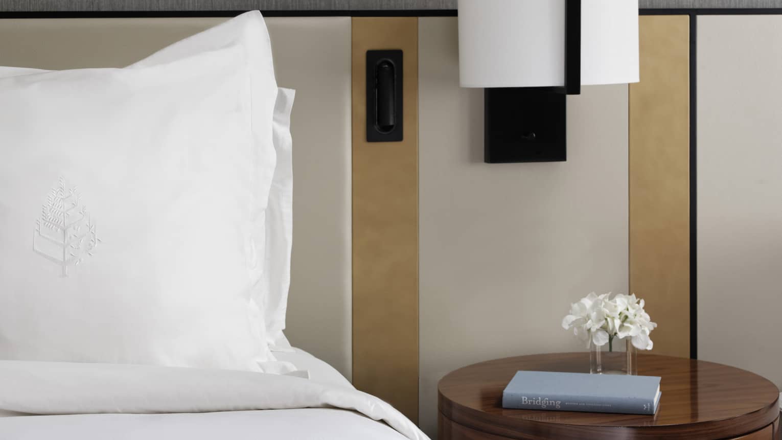 Bed with white linens, round side table with book, reading lamp