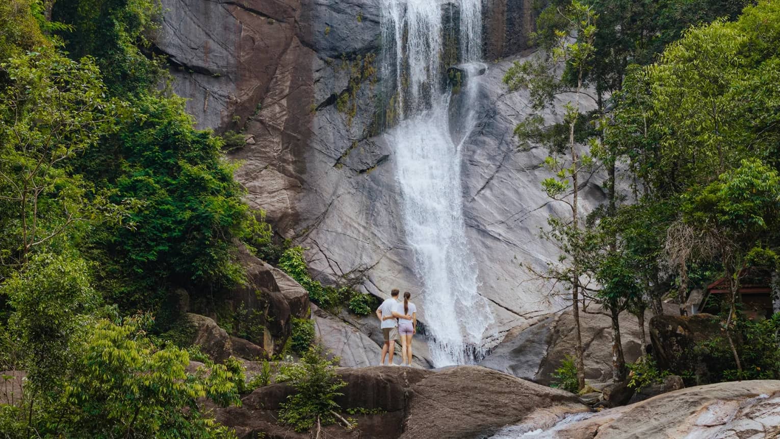 A couple stands at the base of a waterfall surrounded by lush green trees