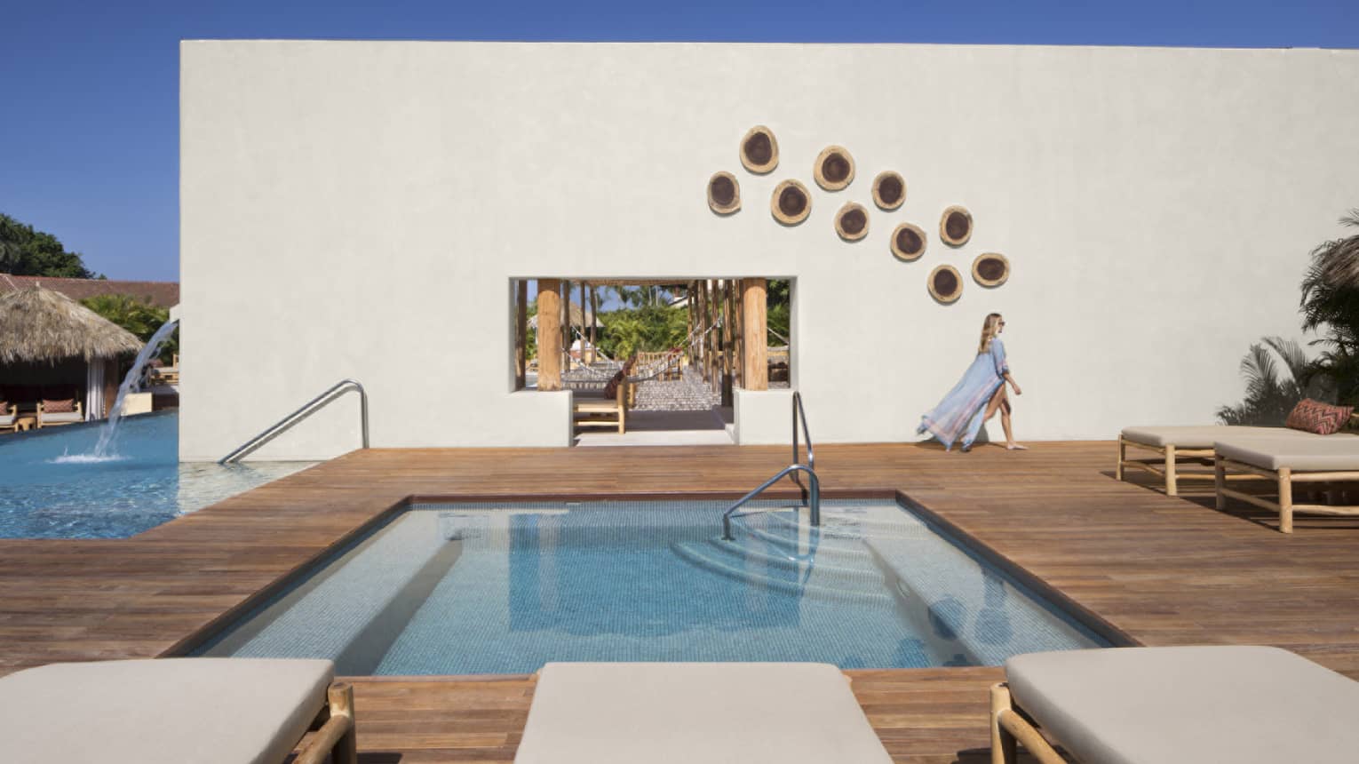 Woman wearing a long, flowy cover-up, walking past a stucco wall on a wooden pool deck