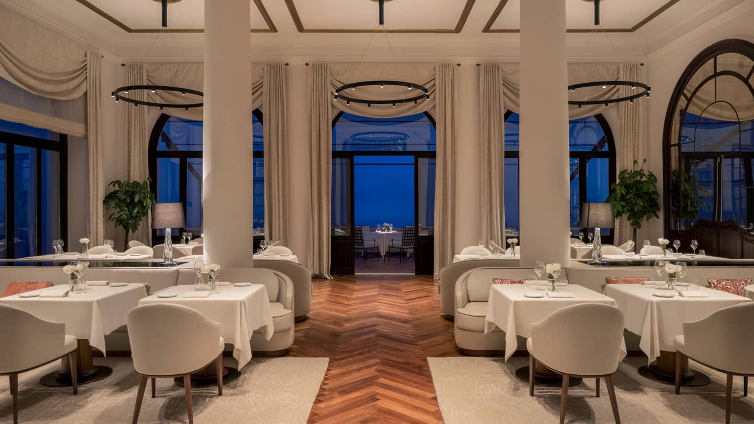 A restaurant with columns, large windows and white cloth tables.