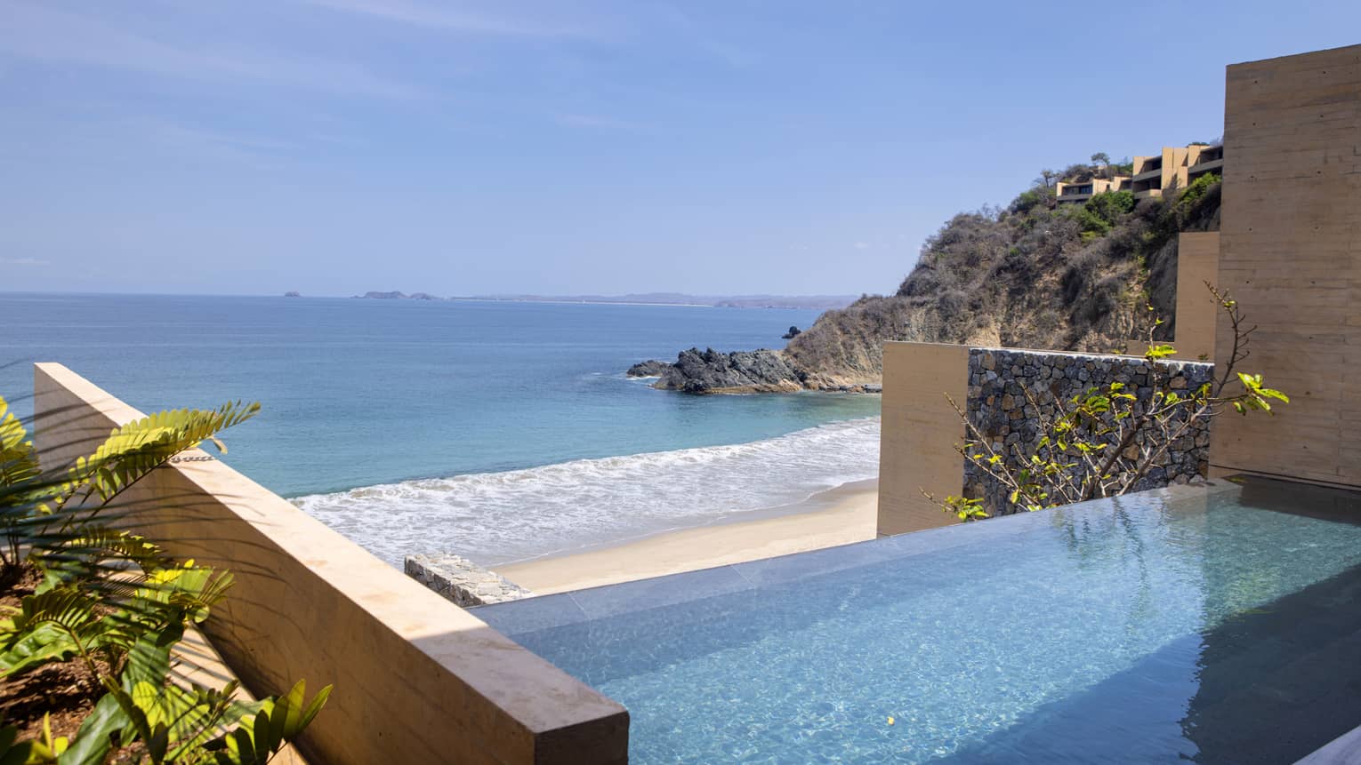 Infinity pool overlooking a pristine beach and rocky coastline, with distant islands visible on the horizon under a clear blue sky. Lush greenery lines the poolside.