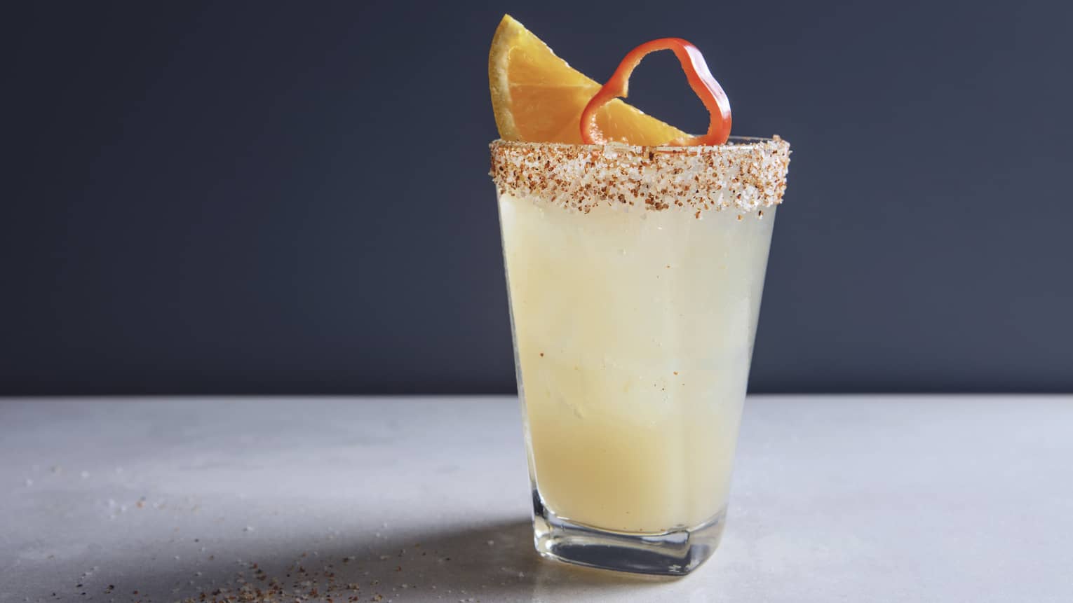 Iced yellow cocktail with a seasoned salt rim, garnished with a slice of lemon and red chili pepper