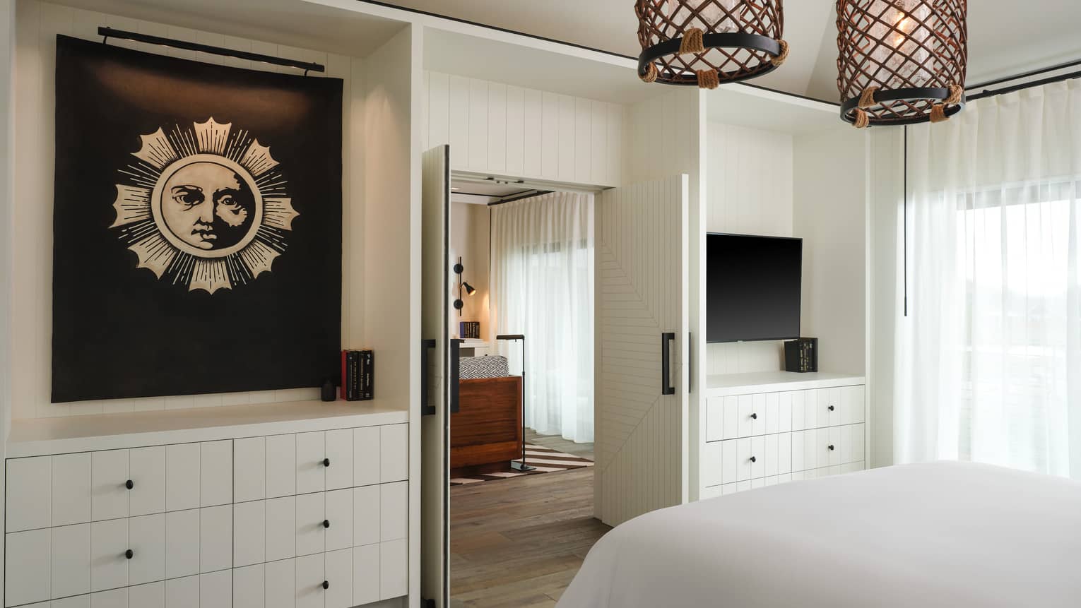 Luxury resort suite bedroom with white cabinetry, sun-themed artwork, wicker pendant lights and a view through double doors into an adjoining room