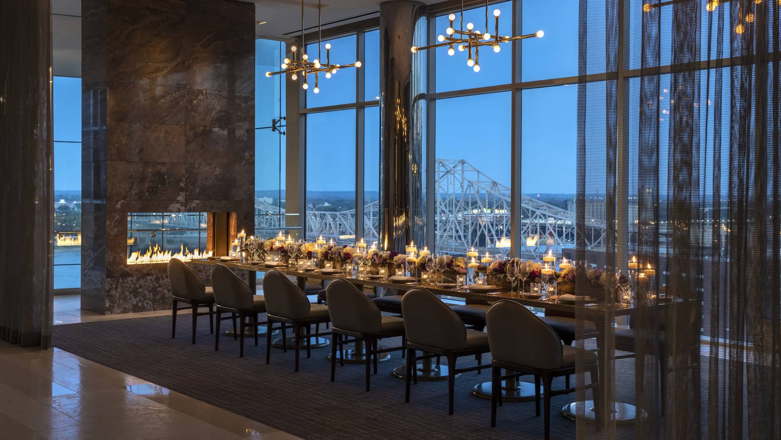 A large dining room with large windows, lights, candles and a large rectangular table.