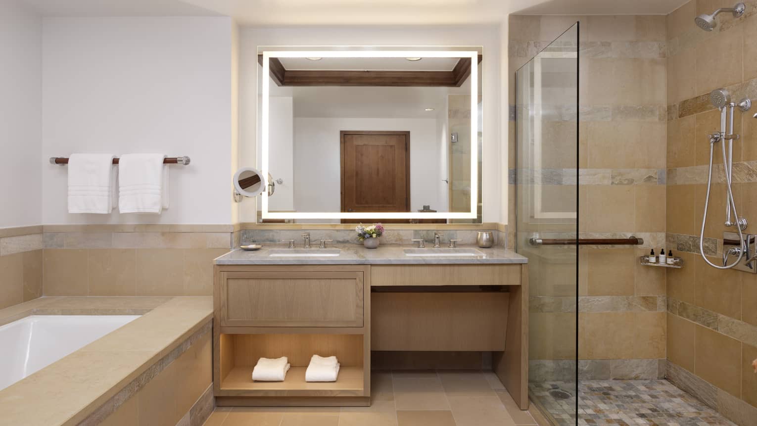 Luxury resort bathroom with a double vanity, backlit mirror, glass-enclosed shower and beige tiled bathtub with towel rack