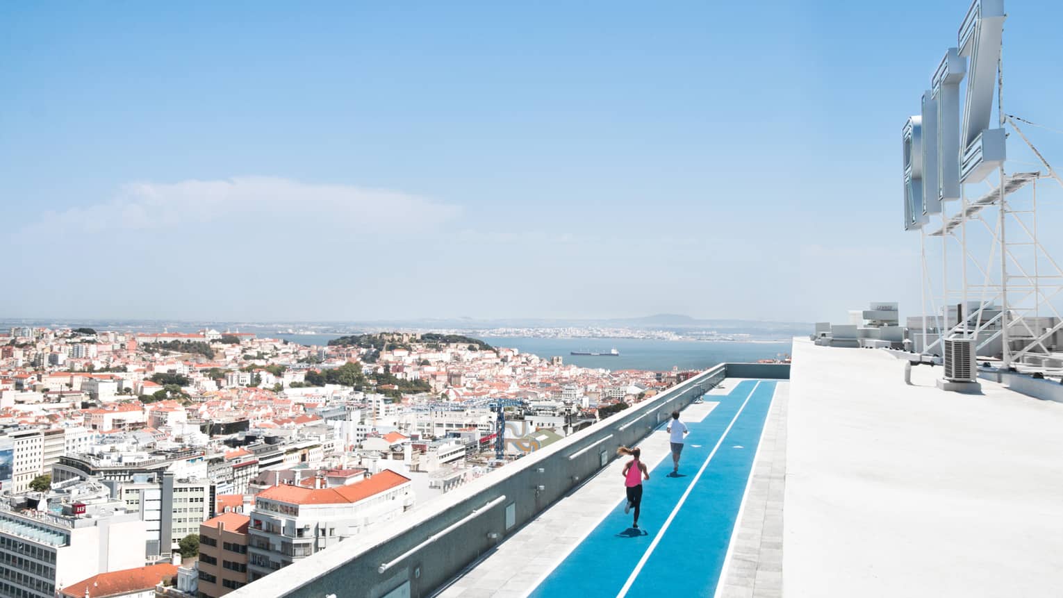 Aerial view of backs of man and woman running on blue rooftop outdoor track, Lisbon city roofs below