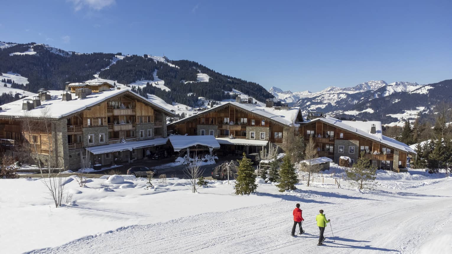 Two people show shoeing on snow-covered path in front of Four Seasons Hotel Megeve with Alps in backdrop