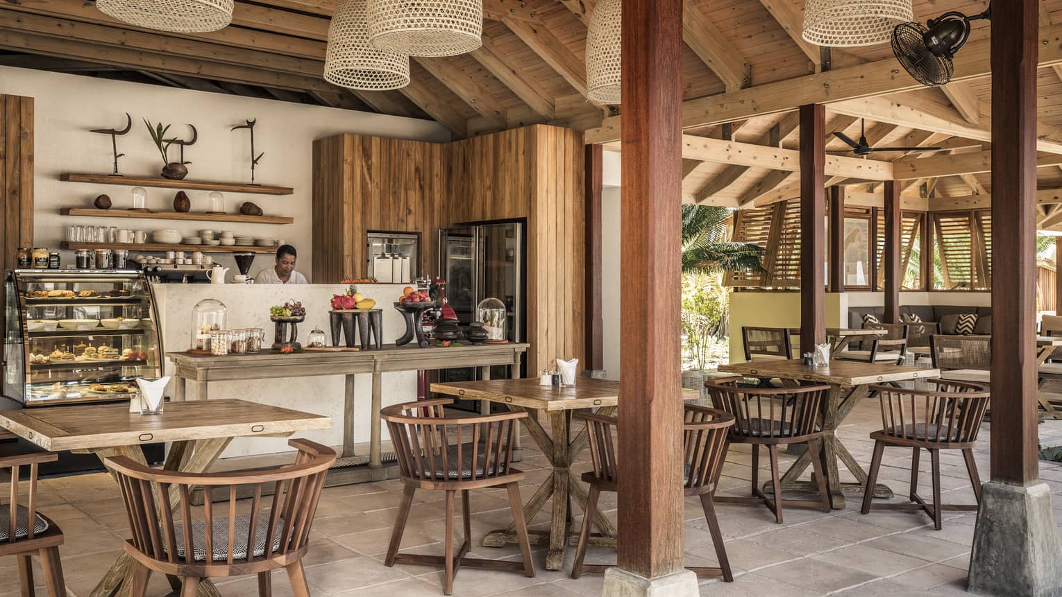Cafe interior with beamed ceilings, wooden dining tables for four, self-service counter, pastry case and staff member in kitchen
