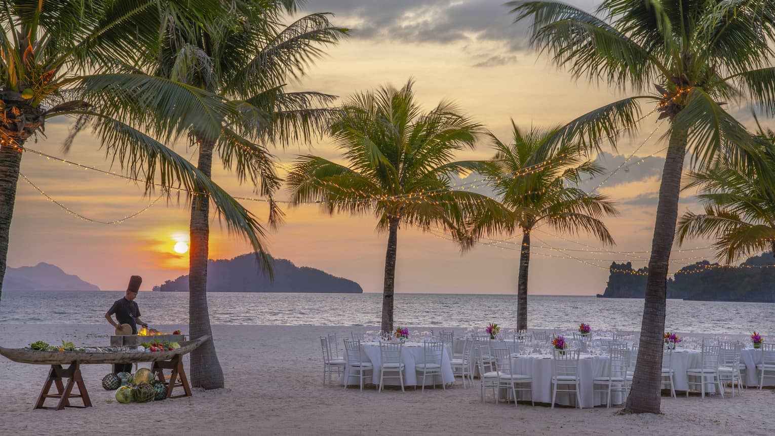 Tables set for dinner on the beach under a canopy of palm trees, a chef cooking over an open flame against the setting sun.