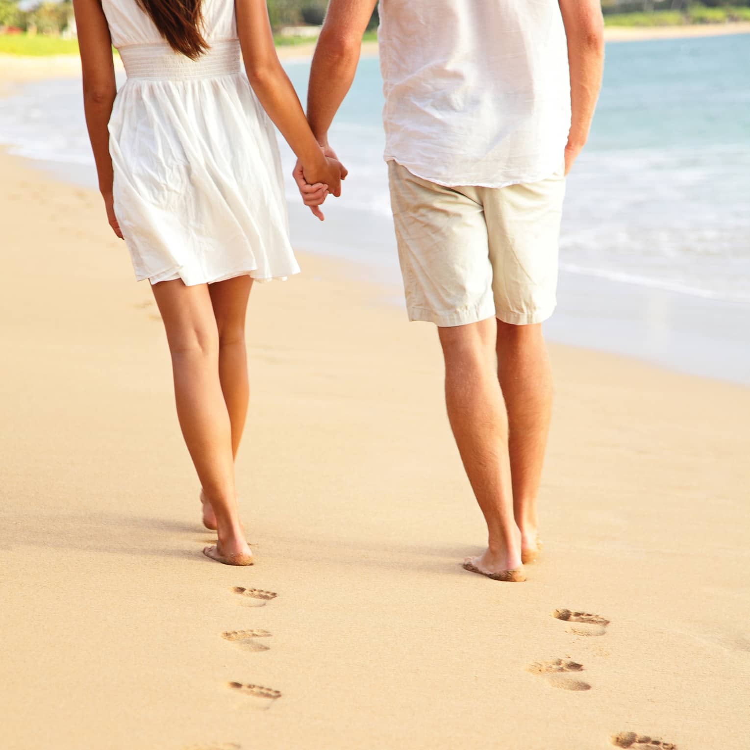 A close-up of a couple holding hands and leaving a trail of footprints as they stroll down a beach near the water’s edge.