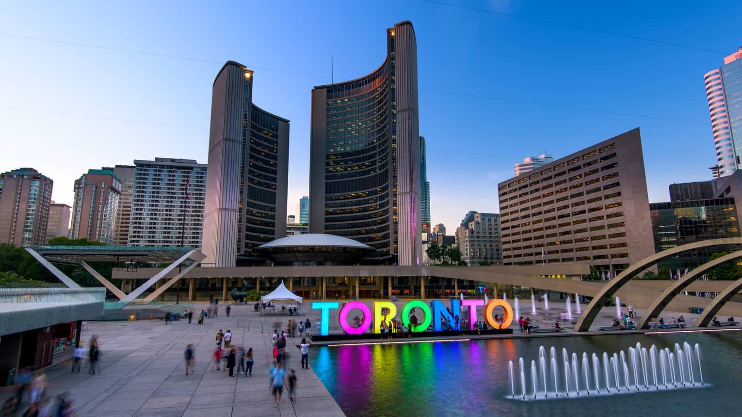 Colourful glowing letters spelling Toronto by fountain people in courtyard under curved City Hall buildings