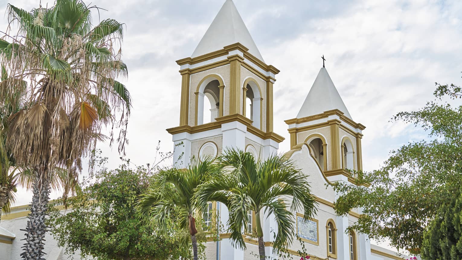 Two church bell towers rise behind palm trees and in front of a cloudy sky. They feature arched windows and gold paint.