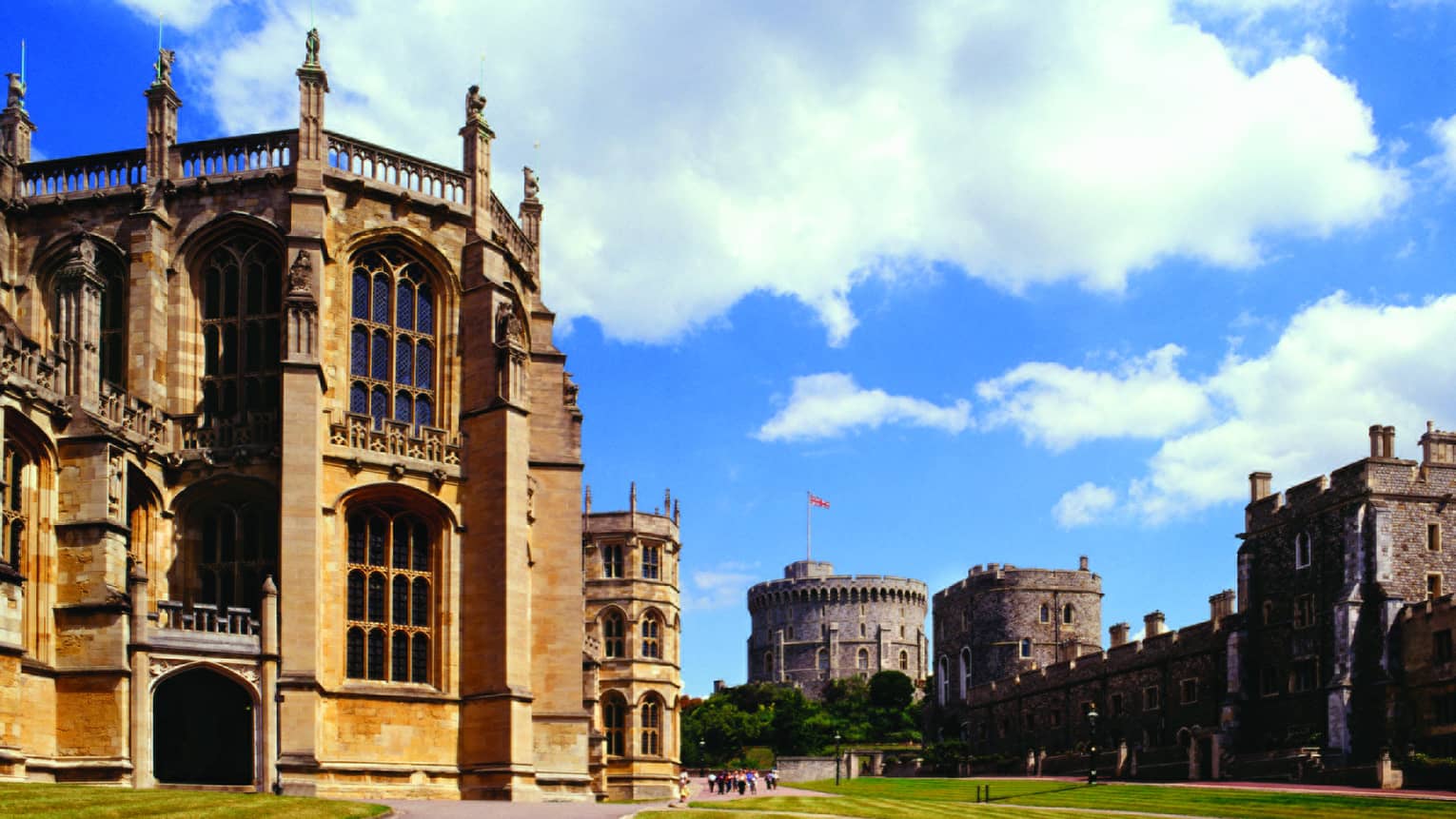 A wide shot of the Windsor Castle, with a manicured lawn, clear sky with large white coulds and a flag blowing in the background