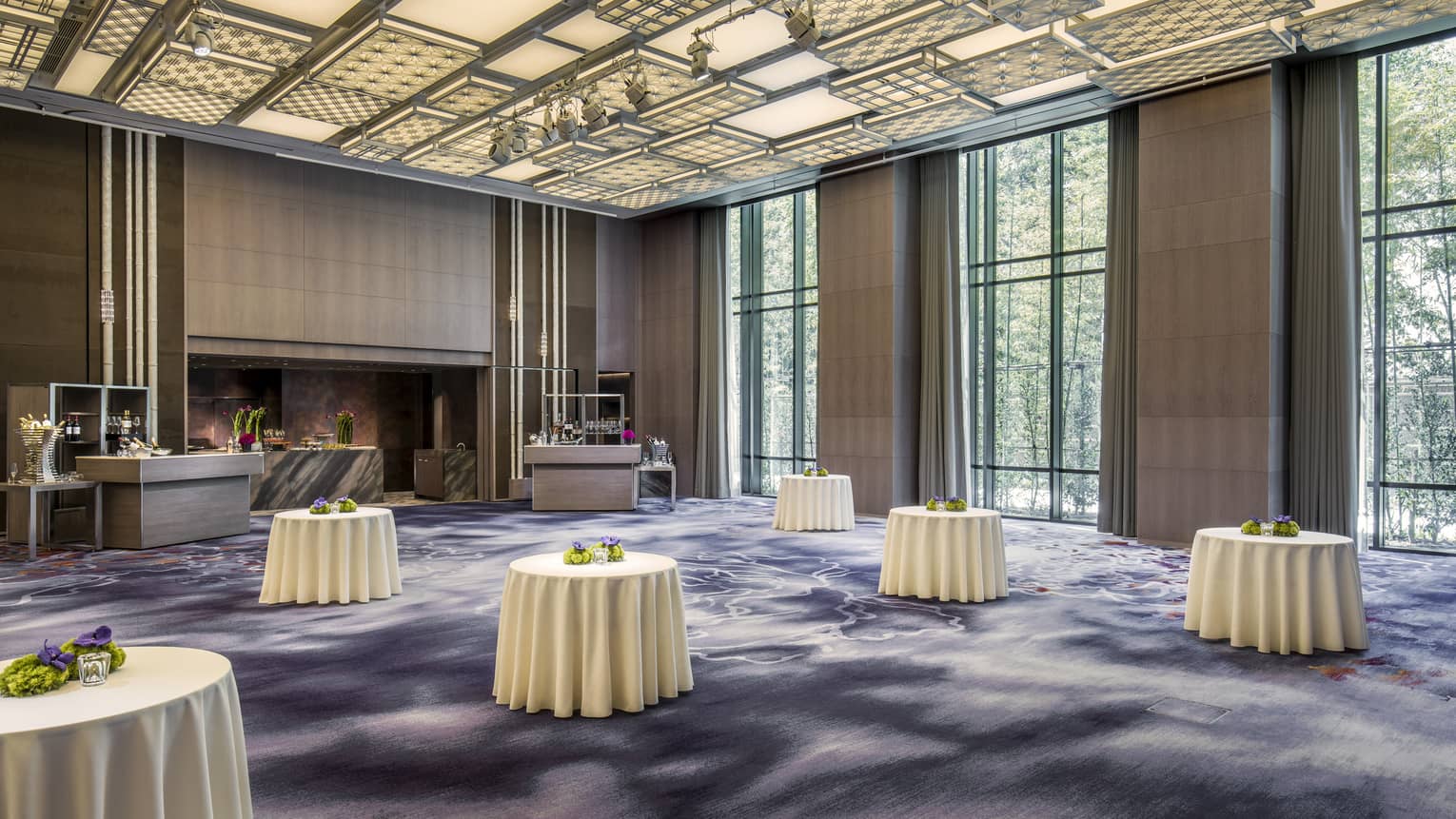 Cocktail reception tables in large ballroom with decorative carpets, ceiling and tall windows