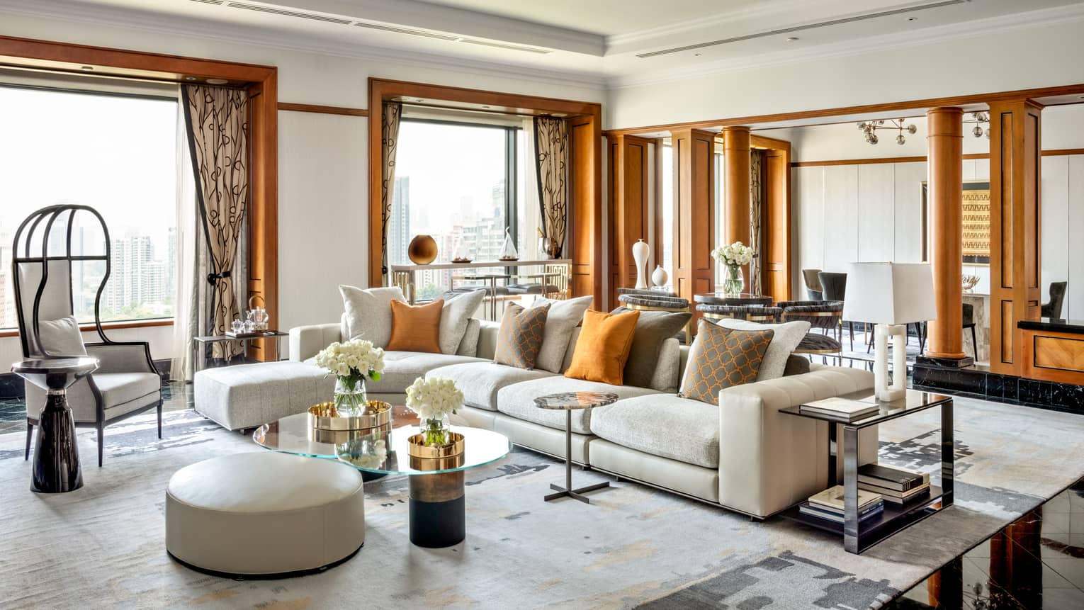 A spacious, luxurious living room with large sectional sofa, orange accent pillows, modern chairs and glass-topped tables. The room features wooden accents and natural light.
