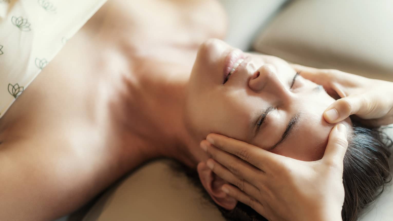 Spa facial, two hands rest on woman's forehead as she closes her eyes, lays under sheet on treatment table
