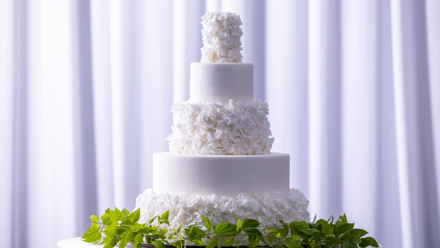 A white floral wedding cake on a stand surrounded by green vines