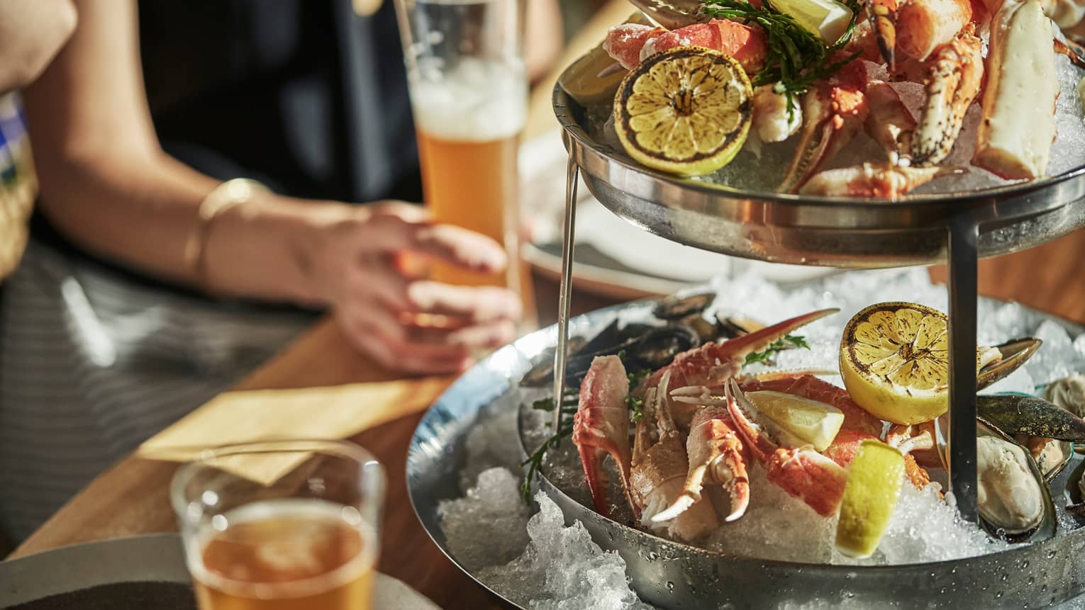 Fish House Tower fresh seafood, crab legs, oysters on two-tiered ice platter, woman's arm holding beer glass