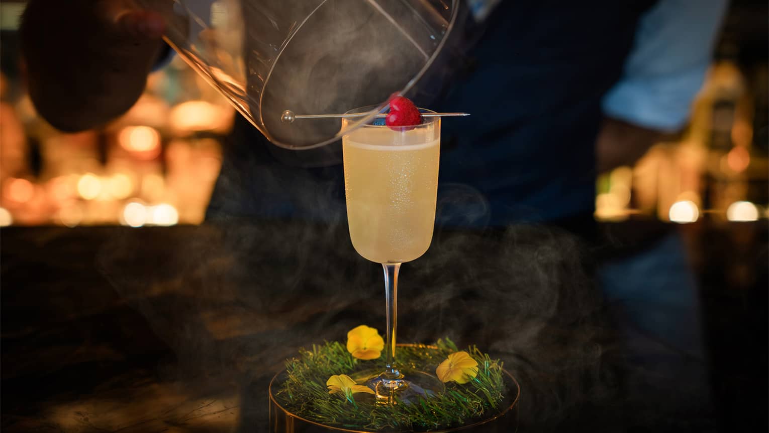 Waiter lifts smoke-filled cloche off of yellow-hued cocktail in stemmed glass