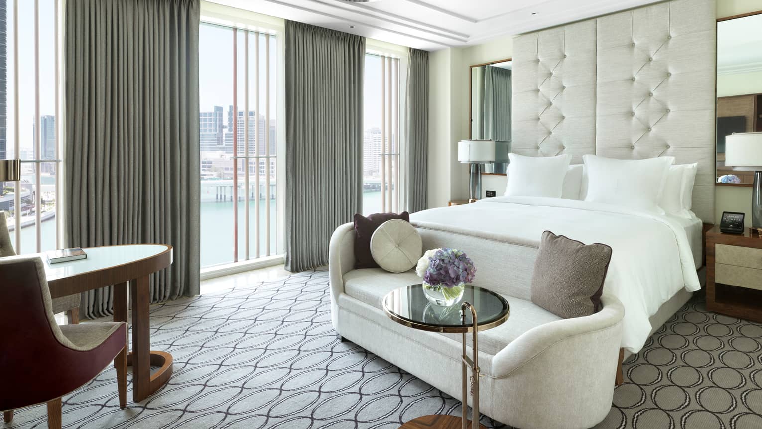 Deluxe room plush white loveseat at foot of bed by sunny window with grey curtains, table and chair