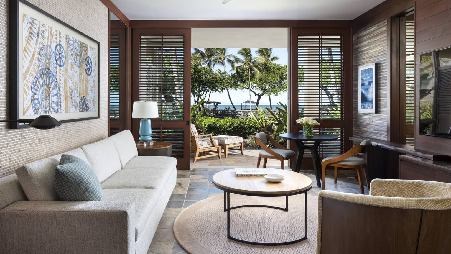 Living room with white sofa, circular coffee table, wall art, dark wooden shutter doors opening to lanai