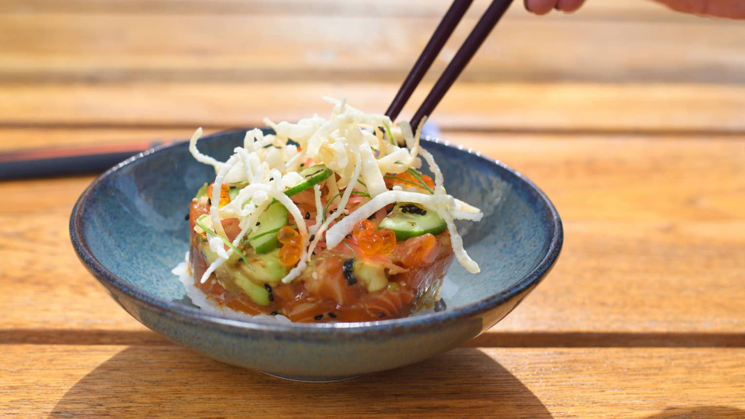 Close-up of chopsticks digging into fresh salmon tartare with crispy garnish in blue bowl