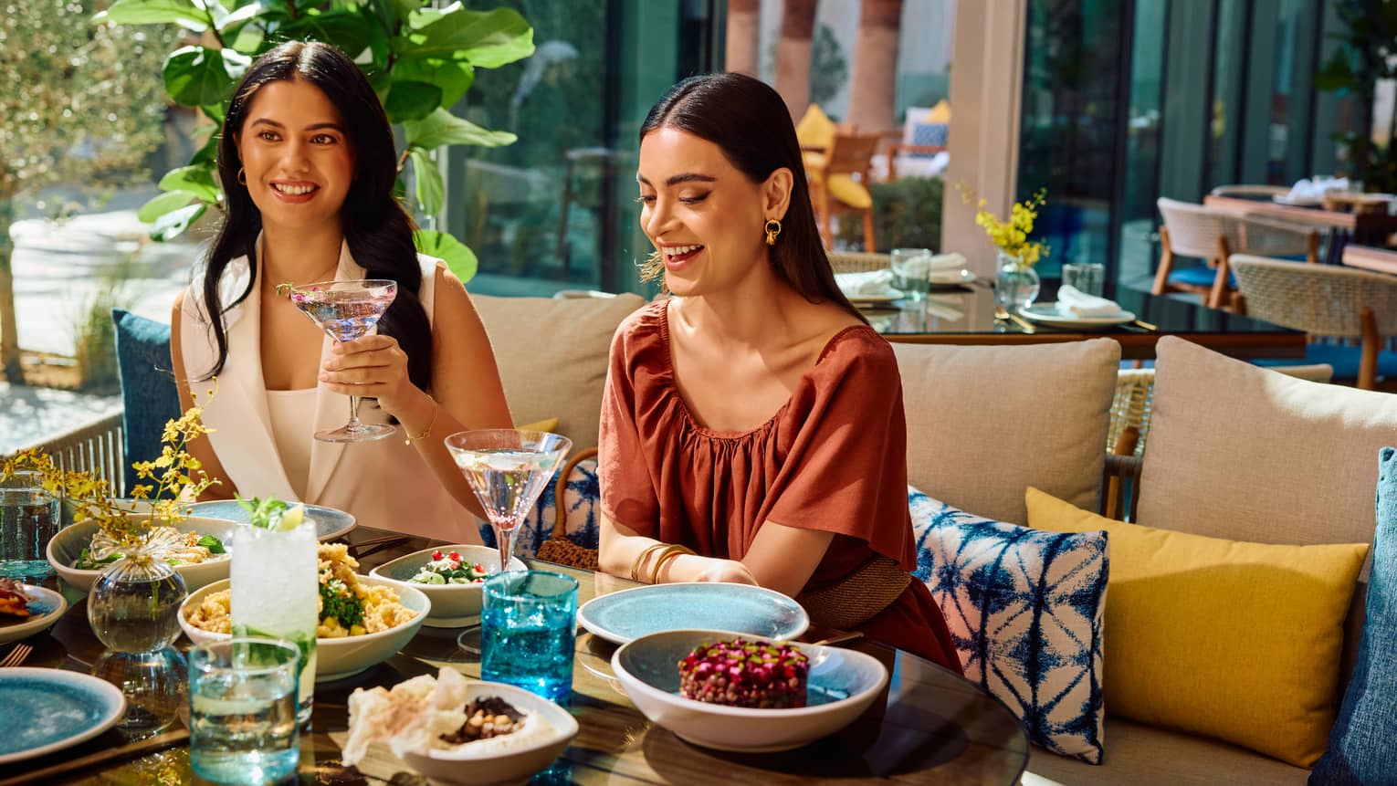 Two guests sit on a sofa filled with yellow and blue throw pillows set next to a table filled with food on an outdoor restaurant patio