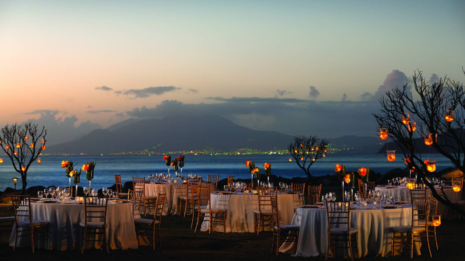 Small dining tables with white linens, trees with lights by ocean at sunset