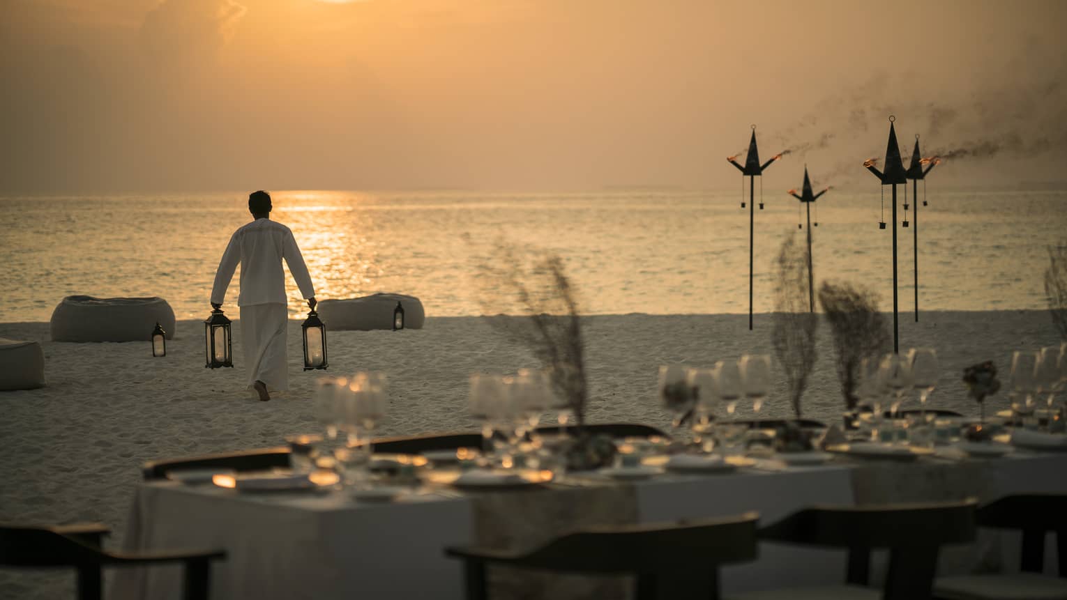 A banquet table is set on the beach as a four seasons member carries two lanterns toward the ocean as the sunsets