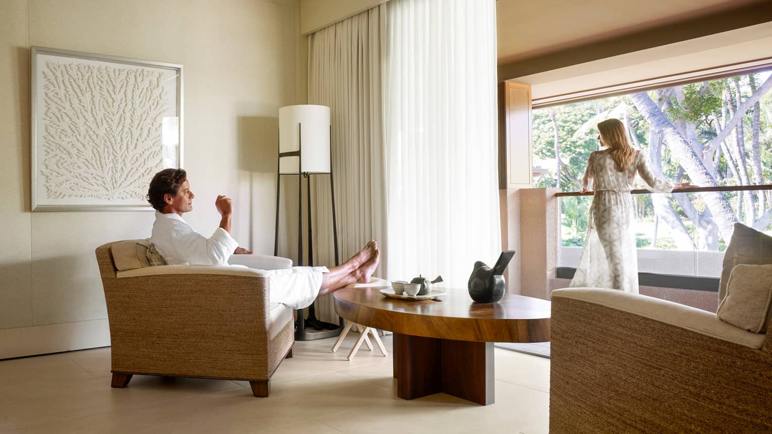 Man in white bathrobe kicks back in armchair in Spa room, woman in sheer robe stands at sunny window