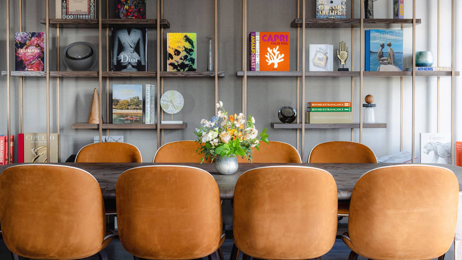 Rectangular table with floral centerpiece and eight suede chairs, copper bookshelf with books and trinkets