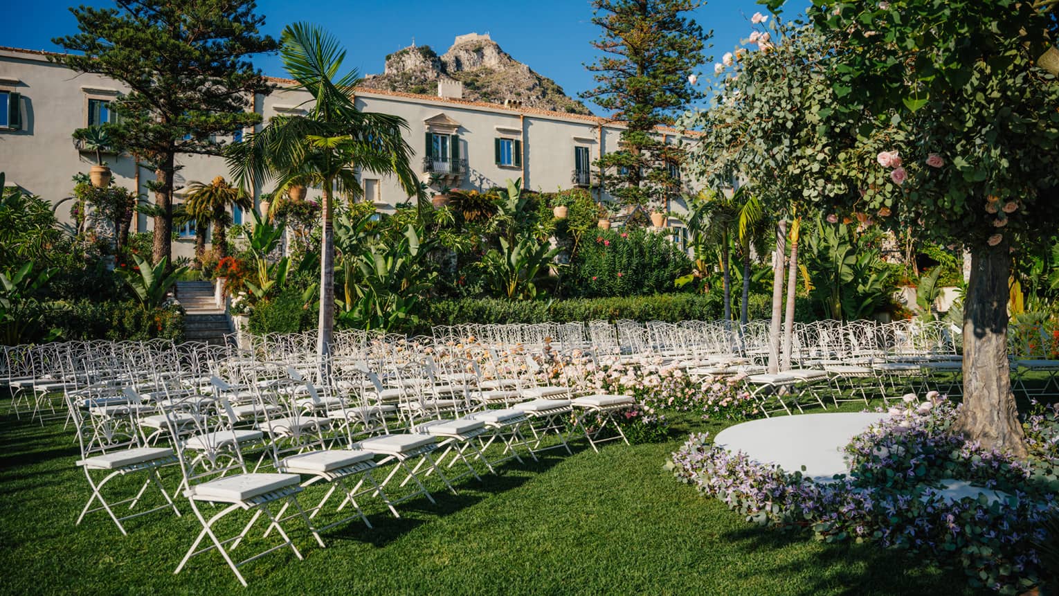 An outdoor event venue on a verdant lawn dotted with trees. Rows of white chairs are arranged in curved lines in front of a round dais framed by flowers.