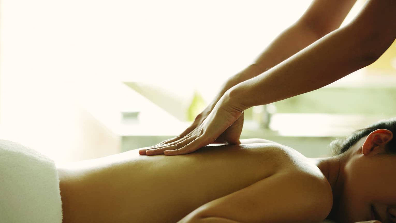 Masseuse reaches over, massages woman's bare back as she lies on spa table