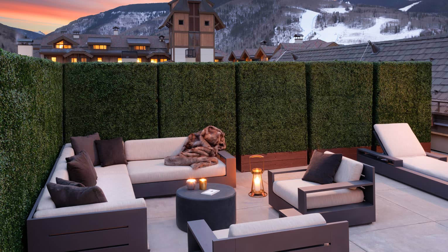 An outdoor seating area with. candles and white sofas near a cabin like village and mountains.