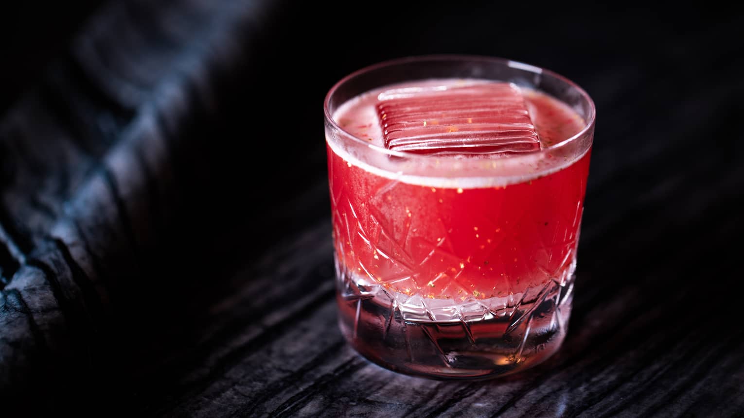 Bright red cocktail in a rocks glass with a large single ice cube, set on a black table