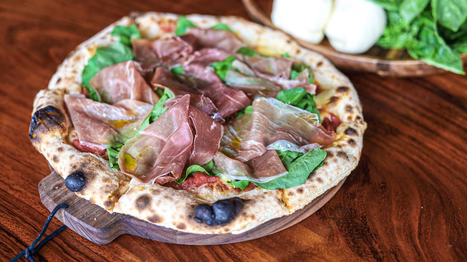 A flatbread on a wooden dish made with mozzarella cheese, tomato sauce, prosciutto di Parma, arugula and olive oil
