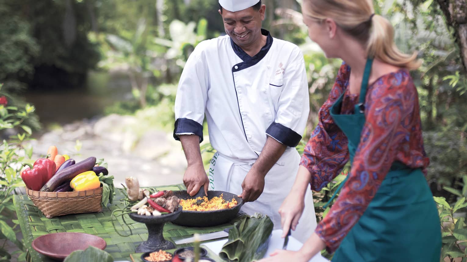 Outdoor cooking class, chef holds pan with rice dish as woman chops vegetables 