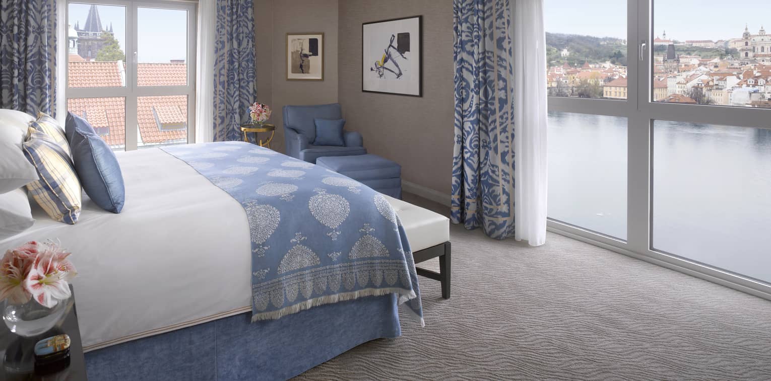Premier Suite bed with blue blanket, pillow accents, in front of floor-to-ceiling corner windows overlooking Vltava River 