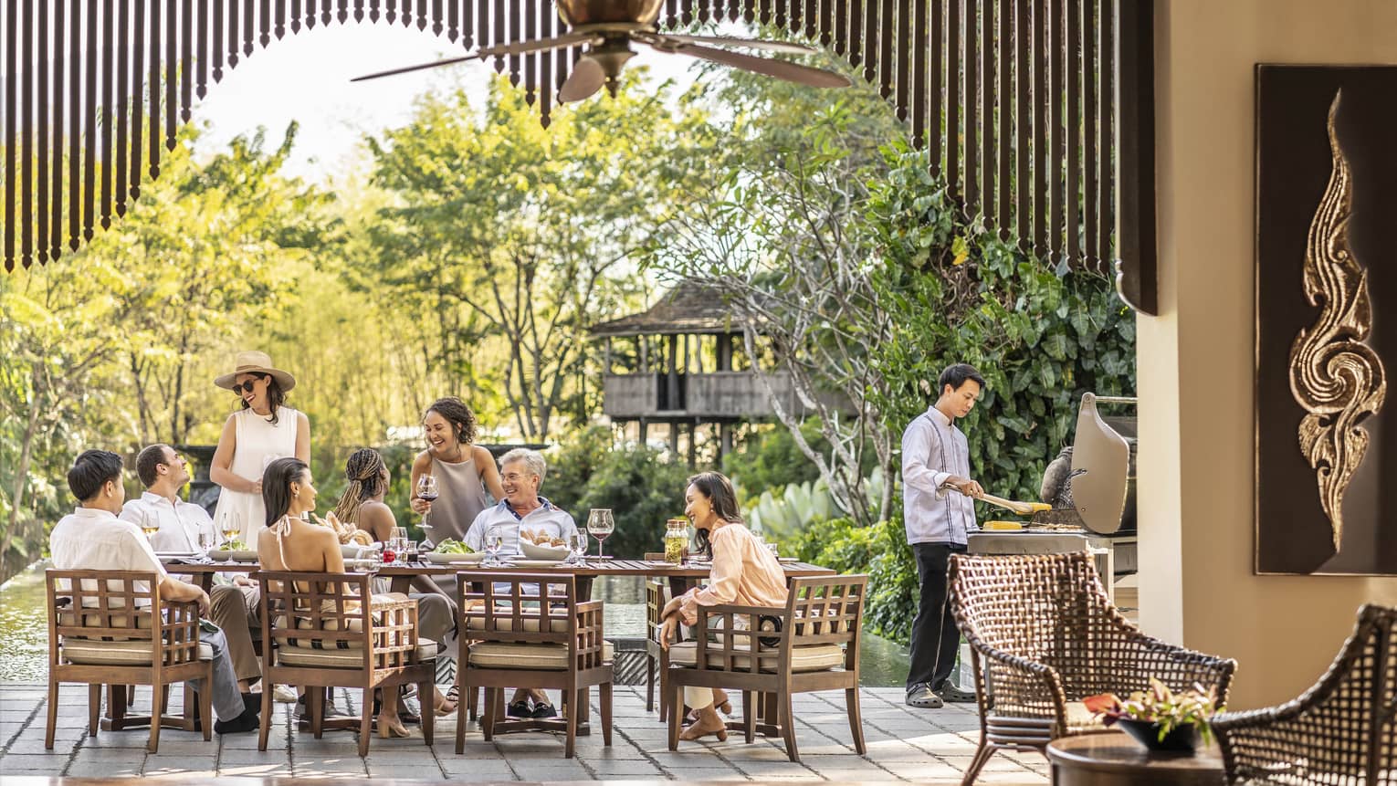A smiling group around a rustic patio set with wine and appetizers, as a chef barbecues corn and light filters through trees.