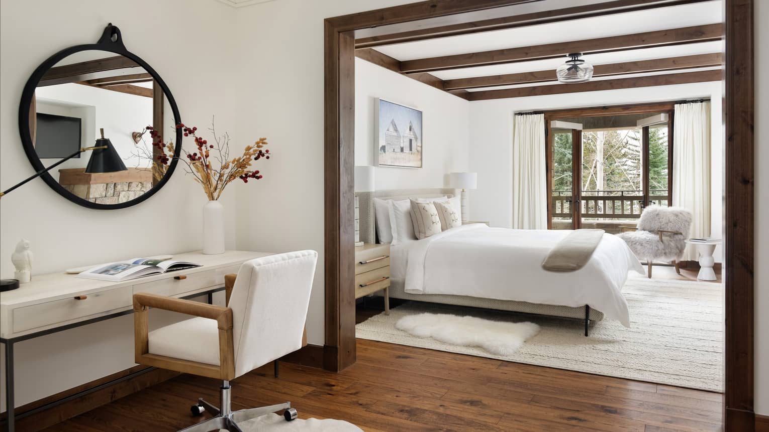 Desk area with chair and round mirror, leading to a connected bedroom with king bed, nightstands with lamps, fluffy armchair and forest-view balcony 