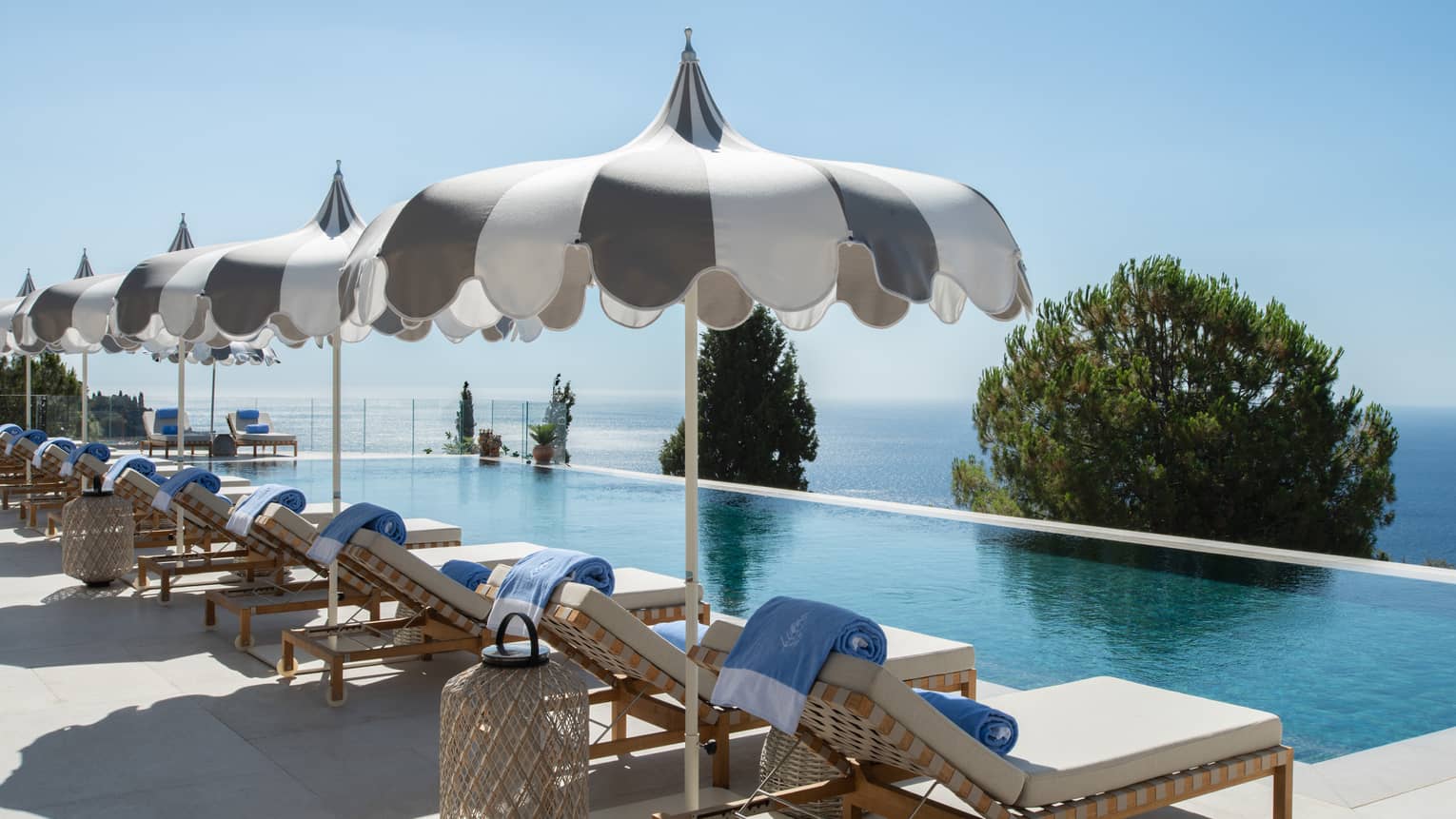 A long inifinity-edge pool with lounge chairs and umbrellas along one side and tree tops and ocean views beyond it