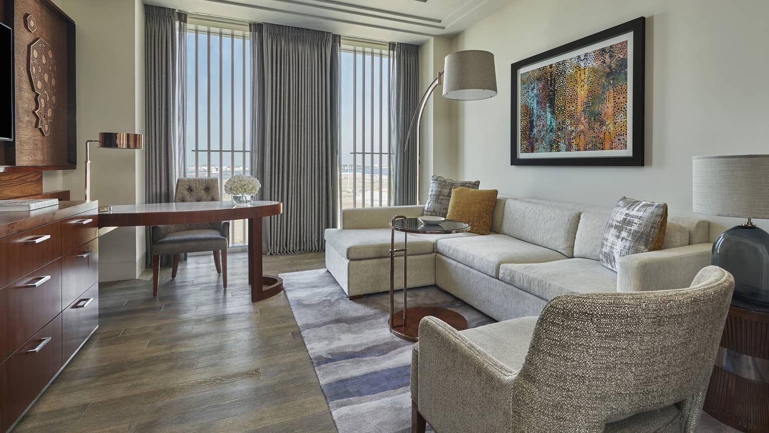 Modern hotel living area with a cream sectional sofa, colorful artwork, a wooden desk and large windows with gray curtains overlooking a scenic view
